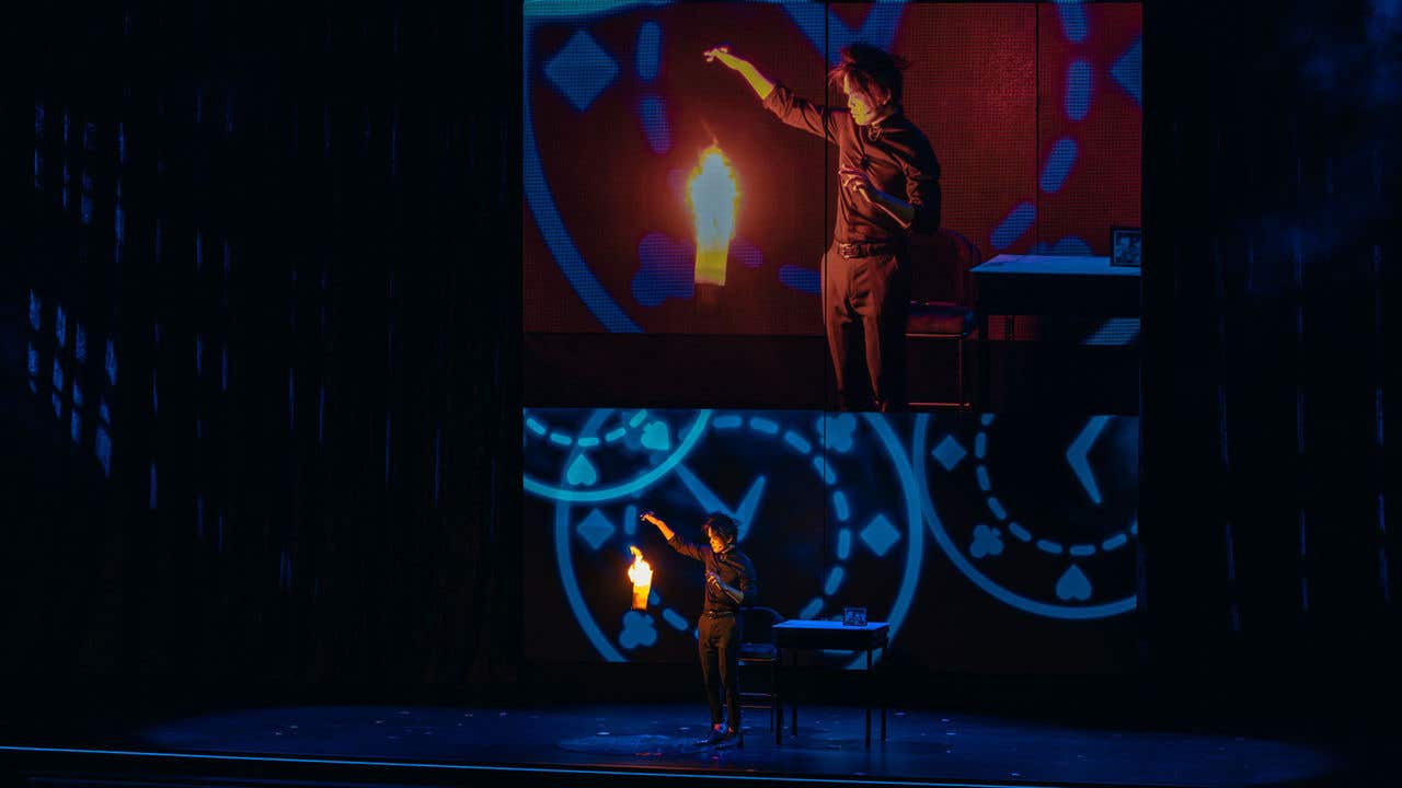 Shin Lim on stage with a fiery torch, projected on a large screen with blue abstract patterns.
